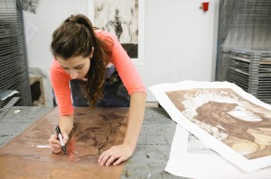 路易斯安那州立大学一名身穿粉色衬衫的版画学生在一块棕色的盘子上雕刻出一个图案